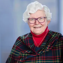 Carolyn is wearing her Clan tartan shawl and a red turtleneck sweater. she has wavy white hair and is smiling